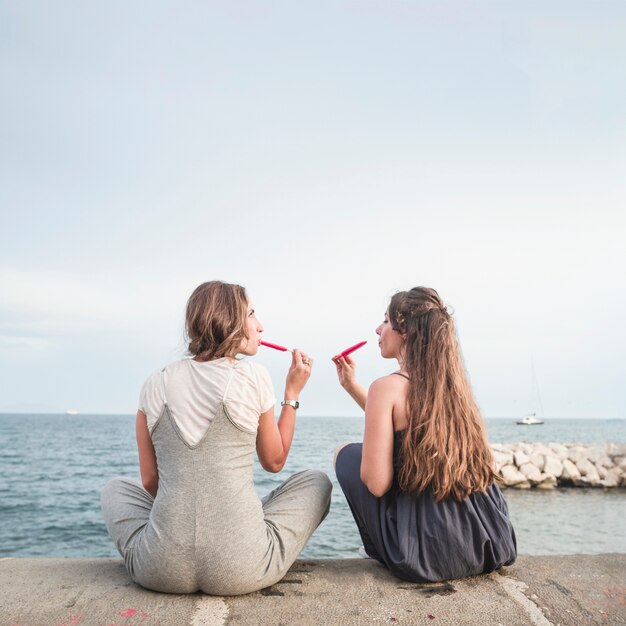 Vista posteriore di due amici femminili seduti sul molo mangiare ghiaccioli rossi