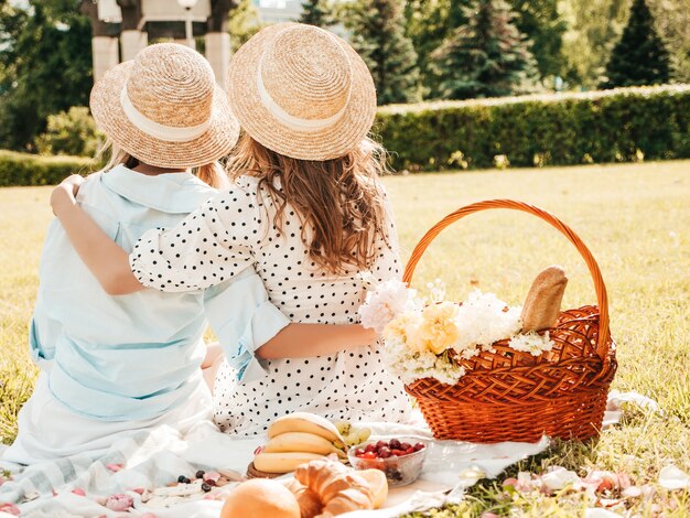 Vista posteriore di due giovani belle ragazze sorridenti hipster in prendisole e cappelli estivi alla moda. Donne che fanno picnic all'esterno.