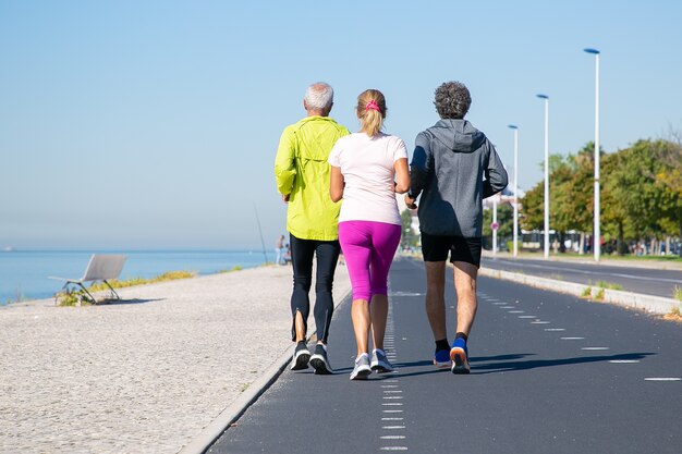 Vista posteriore di pareggiatori maturi in abiti sportivi in esecuzione sulla pista lungo la riva del fiume. Lunghezza intera, copia dello spazio. Attività o concetto di stile di vita sano