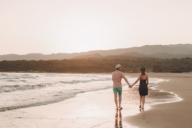 Vista posteriore di una coppia romantica hipster camminando sulla spiaggia durante le vacanze estive al tramonto