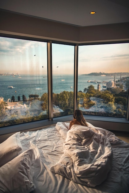 Vista posteriore di una donna sdraiata sul letto guardando la spiaggia attraverso le finestre di vetro
