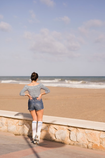 Vista posteriore di una femmina pattinatore con la mano sulla sua anca guardando il mare