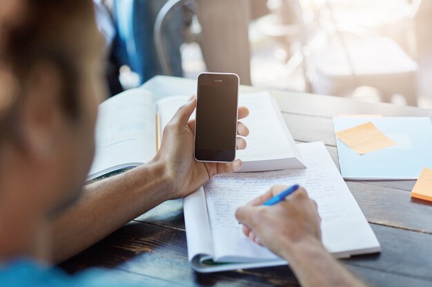 Vista posteriore di uno studente di carnagione scura in possesso di smart phone con schermo vuoto per le tue informazioni, annotare le note nel quaderno mentre studia nella biblioteca o mensa universitaria