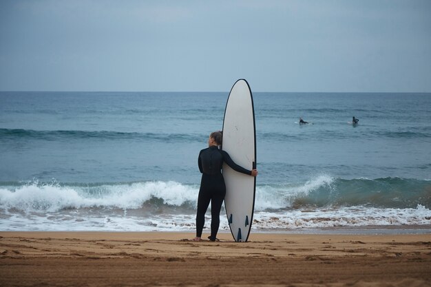 Vista posteriore sulla bella giovane ragazza di surf che abbraccia il suo longboard sulla riva dell'oceano e guardare le onde prima del surf