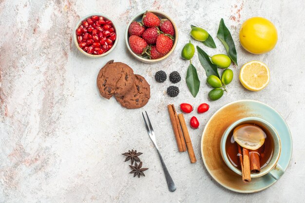 Vista ravvicinata dall'alto biscotti al cioccolato biscotti al cioccolato una tazza di tè con limone e bastoncini di cannella forchetta ciotole di frutti di bosco agrumi melograno sul tavolo