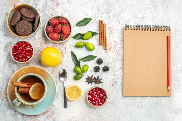 Vista ravvicinata dall'alto biscotti e frutti di bosco taccuino crema e matita rossa accanto alla cannella i biscotti appetitosi fragole cucchiaio di limone una tazza di tè agrumi sul tavolo