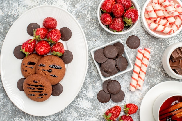 Vista ravvicinata dall'alto biscotti fragole e cioccolatini rotondi sul piatto ovale ciotole di caramelle fragole cioccolatini e tè alla cannella sul tavolo grigio-bianco