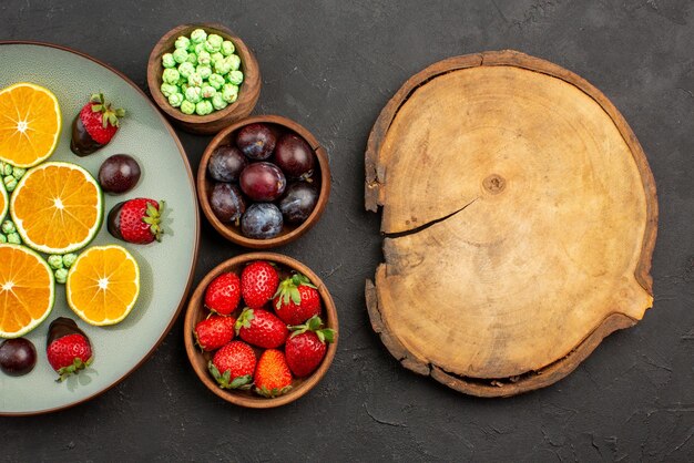 Vista ravvicinata dall'alto frutta ricoperta di cioccolato caramelle verdi alla fragola ricoperte di cioccolato arancia tritata e ciotole di frutti diversi bacche e dolci accanto alla tavola da cucina in legno