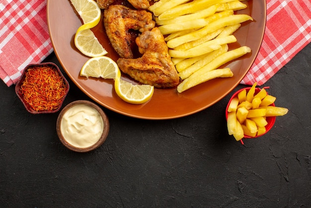 Vista ravvicinata dall'alto gustoso pollo appetitose ali di pollo patatine fritte e ciotole al limone di diversi tipi di salse e spezie al centro del tavolo scuro