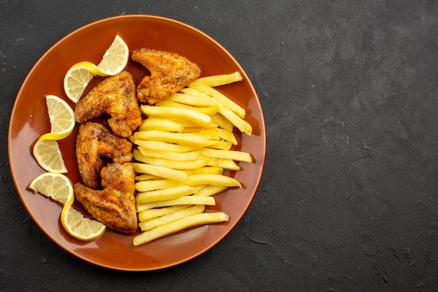Vista ravvicinata dall'alto piatto arancione fastfood di ali di pollo con patatine fritte e limone sul lato sinistro del tavolo scuro
