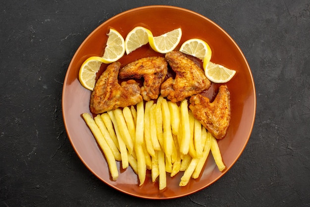 Vista ravvicinata dall'alto piatto arancione fastfood di ali di pollo con patatine fritte e limone sul tavolo scuro