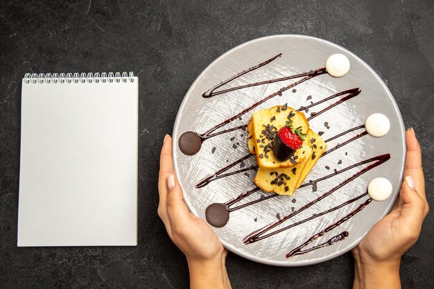 Vista ravvicinata dall'alto piatto di torta con fragole ricoperte di cioccolato e salsa di cioccolato nelle mani accanto al taccuino bianco