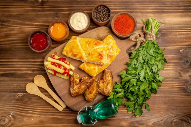 Vista ravvicinata dall'alto spezie salse ali di pollo patatine fritte con ketchup e torta sulla tavola della cucina accanto alle ciotole di spezie colorate e salse cucchiai di legno erbe e bottiglia