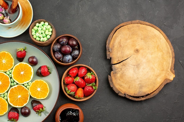 Vista ravvicinata dall'alto tazza di tè e frutta una tazza di tè ricoperte di cioccolato fragole arancioni tritate caramelle verdi e ciotole di diverse bacche e dolci accanto al tagliere