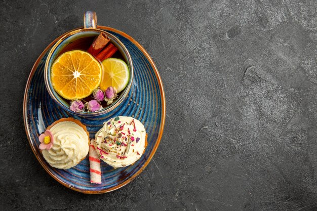 Vista ravvicinata dall'alto una tazza di tè al limone un piattino blu degli appetitosi cupcakes con crema e una tazza di tisana con limone e cannella sul tavolo scuro