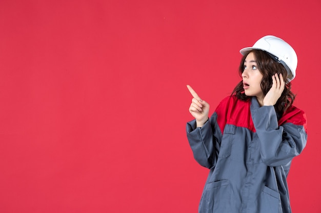 Vista ravvicinata del costruttore femminile scioccato in uniforme con elmetto e indicando qualcosa sul lato destro sul muro rosso isolato