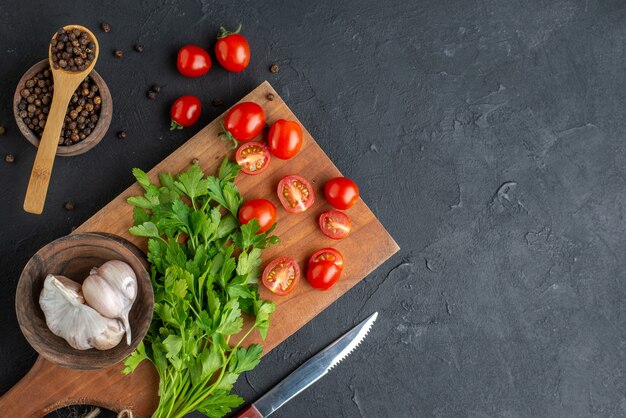 Vista ravvicinata del fascio verde fresco intero di pomodori tagliati aglio su tagliere di legno coltello pepe su superficie nera angosciata