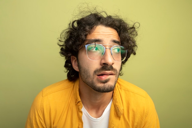 Vista ravvicinata del giovane uomo bello ansioso con gli occhiali guardando il labbro mordace laterale isolato sulla parete verde oliva