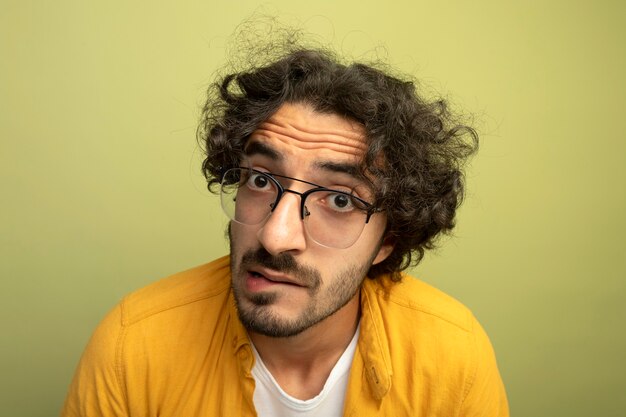 Vista ravvicinata del giovane uomo bello colpito con gli occhiali guardando il labbro mordace anteriore isolato sulla parete verde oliva