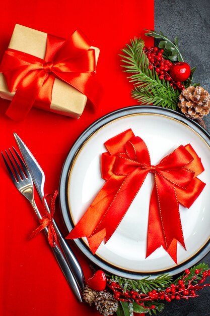 Vista ravvicinata del nuovo anno sfondo con nastro rosso sulla piastra della cena set di posate decorazione accessori rami di abete accanto a un regalo su un tovagliolo rosso