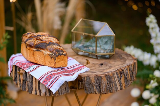 Vista ravvicinata del pane cotto su un asciugamano da ricamo posizionato su un tavolo di legno vicino a una scatola di vetro preparata per la cerimonia di matrimonio all'aperto