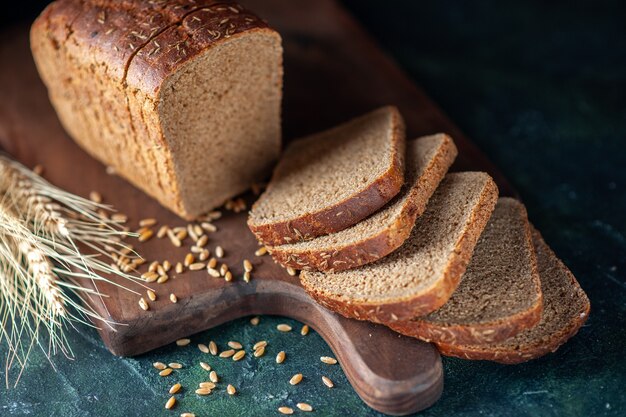 Vista ravvicinata del pane nero dietetico con spighe di grano su tagliere di legno su sfondo blu scuro colori