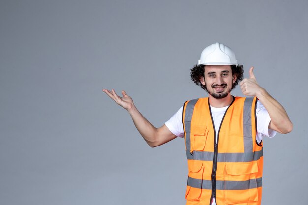 Vista ravvicinata dell'architetto maschio sorridente in giubbotto di avvertimento con casco di sicurezza e indicando qualcosa sul lato destro facendo gesto ok sulla parete grigia dell'onda