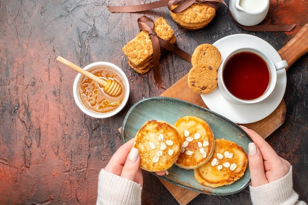 Vista ravvicinata della mano che prende il vassoio con frittelle fresche una tazza di tè nero su un tagliere di legno biscotti impilati miele latte su una superficie scura