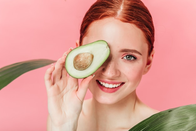 Vista ravvicinata della splendida donna allo zenzero con avocado. Studio shot di eccitata ragazza caucasica con cibo sano isolato su sfondo rosa.