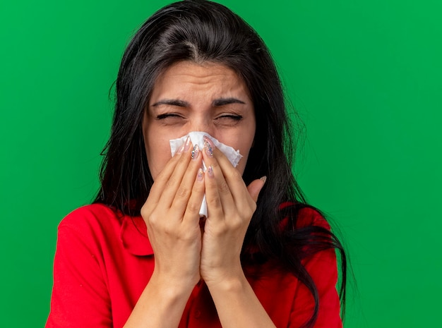 Vista ravvicinata di accigliata giovane donna malata pulendosi il naso con il tovagliolo con gli occhi chiusi isolato sulla parete verde