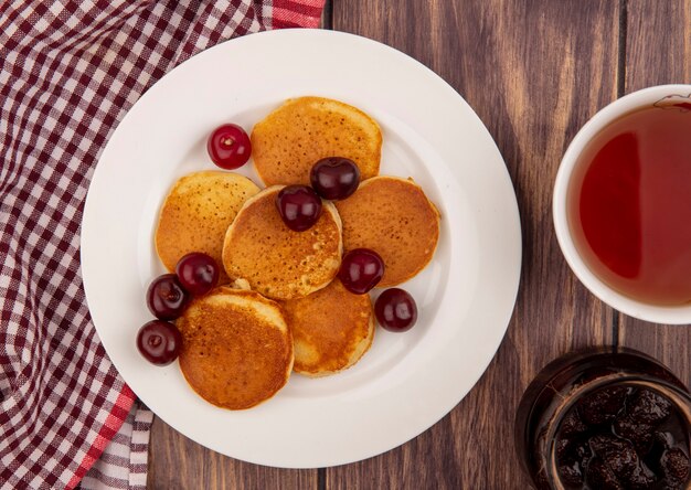 Vista ravvicinata di frittelle con ciliegie in piastra su plaid panno con tazza di tè e marmellata di fragole su sfondo di legno