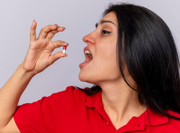 Vista ravvicinata di giovane donna malata che guarda dritto prendendo capsula medica isolata sul muro bianco