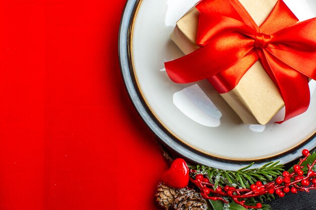 Vista ravvicinata di piatti da pranzo con regalo su di esso e rami di abete con cono di conifera accessorio decorazione su un tovagliolo rosso