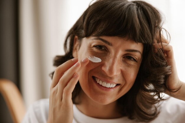 Vista ravvicinata di prodotti donna felice per crema per la pelle guardando lo specchio