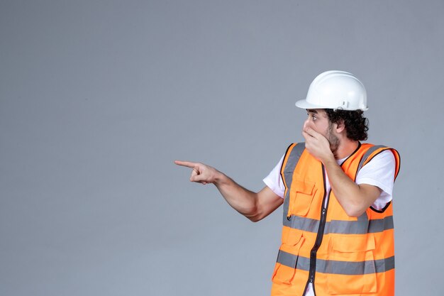 Vista ravvicinata di un curioso architetto maschio in giubbotto di avvertimento con casco di sicurezza e che punta qualcosa sul lato destro sulla parete grigia dell'onda