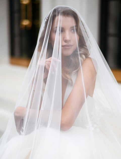 Vista ravvicinata di una donna seria e attraente in un lungo abito bianco che alza la mano nascondendo la faccia sotto il velo e distogliendo lo sguardo mentre si siede sullo sfondo di un edificio moderno