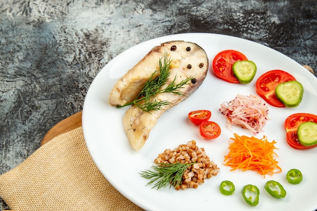 Vista ravvicinata frontale del pasto di grano saraceno di pesce bollito servito con verdure verdi su un piatto bianco su asciugamano nudo su tagliere di legno sulla superficie del ghiaccio