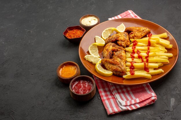 Vista ravvicinata laterale cibo su piatto arancione appetitose patatine fritte ali di pollo con limone e ketchup e ciotole di salse e spezie su tovaglia a scacchi bianco-rosa sulla destra