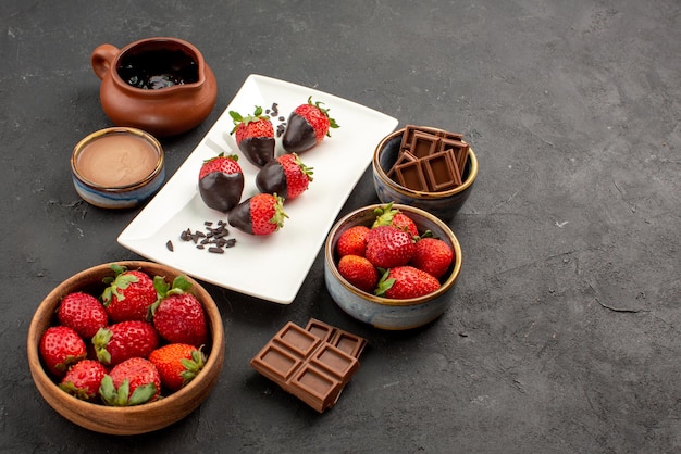 Vista ravvicinata laterale fragole crema al cioccolato piatto bianco di fragole ricoperte di cioccolato barrette di cioccolato e crema di cioccolato in una ciotola sul tavolo scuro