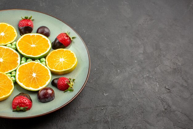 Vista ravvicinata laterale frutti ricoperti di cioccolato appetitose caramelle verdi arancioni tritate e fragole ricoperte di cioccolato sul lato sinistro del tavolo nero