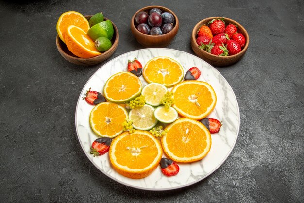 Vista ravvicinata laterale frutti su ciotole da tavola di agrumi e bacche e piatto di fragole e agrumi ricoperti di cioccolato al centro del tavolo scuro