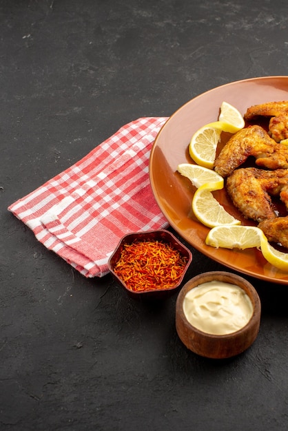 Vista ravvicinata laterale piatto di ali di pollo fast food patatine fritte con limone e ciotole di salse e spezie su tovaglia a scacchi bianco-rosa sul lato destro del tavolo nero