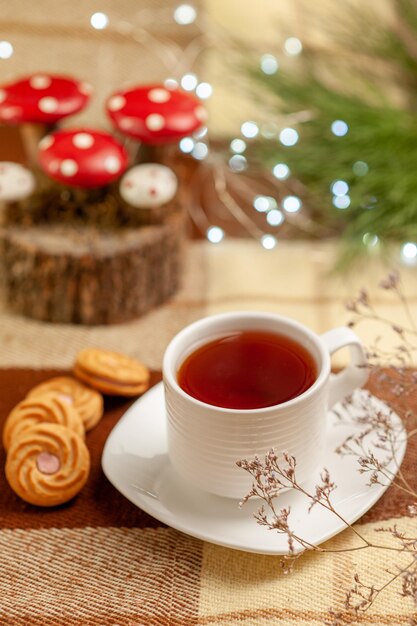 Vista ravvicinata laterale teiera biscotti appetitosi e una tazza di tè su un piattino accanto alla teiera e ai rami degli alberi sulla tovaglia a scacchi