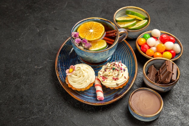 Vista ravvicinata laterale tisana una tazza di tè al limone e cupcakes con crema accanto alle ciotole di cioccolato fette di crema al cioccolato lime e dolci colorati sul tavolo