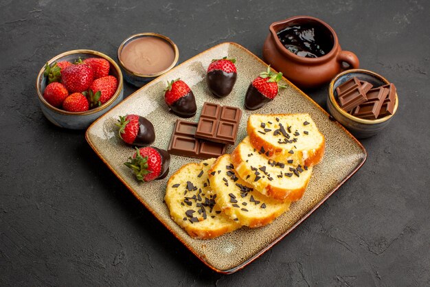 Vista ravvicinata laterale torta con fragole crema al cioccolato fragola e cioccolato in ciotole marroni e piatto di torta con fragole ricoperte di cioccolato su sfondo nero