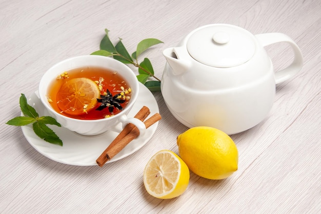 Vista ravvicinata laterale una tazza di tè al limone una tazza di tisana con cannella al limone accanto alla teiera e foglie sul tavolo bianco