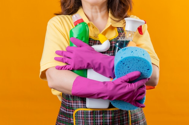 Vista ritagliata della donna che indossa il grembiule e guanti di gomma che tengono gli strumenti per la pulizia in piedi su sfondo arancione isolato