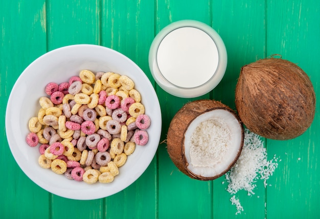 Vista superiore dei cereali multicolori e del ciclo su una ciotola bianca con un bicchiere di latte e una noce di cocco su superficie verde