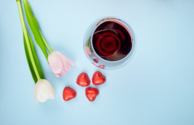 Vista superiore dei fiori bianchi e rosa del tulipano di colore con le caramelle a forma di cuore sparse in stagnola rossa e un bicchiere di vino sulla tavola blu
