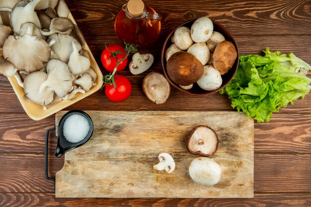 Vista superiore dei funghi freschi in ciotola e pomodori con lattuga e un bordo di legno con sale e funghi affettati su legno rustico